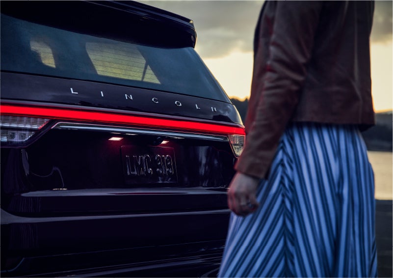 A person is shown near the rear of a 2023 Lincoln Aviator® SUV as the Lincoln Embrace illuminates the rear lights | McLarty Daniel Lincoln in Bentonville AR
