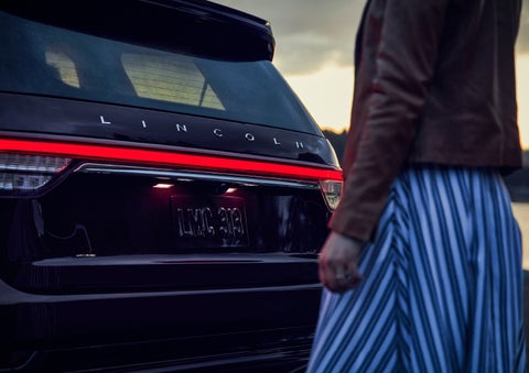 A person is shown near the rear of a 2024 Lincoln Aviator® SUV as the Lincoln Embrace illuminates the rear lights | McLarty Daniel Lincoln in Bentonville AR